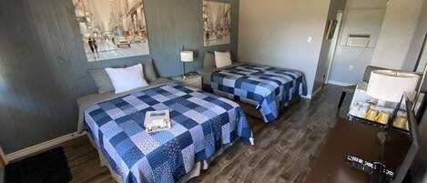 A hotel room with two beds, a desk with a lamp, a small table with toiletries, and artwork on the walls.
