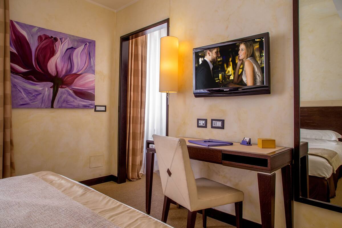 Een hotelkamer met een bed, een bureau met stoel, een televisie en een bloem schilderij.