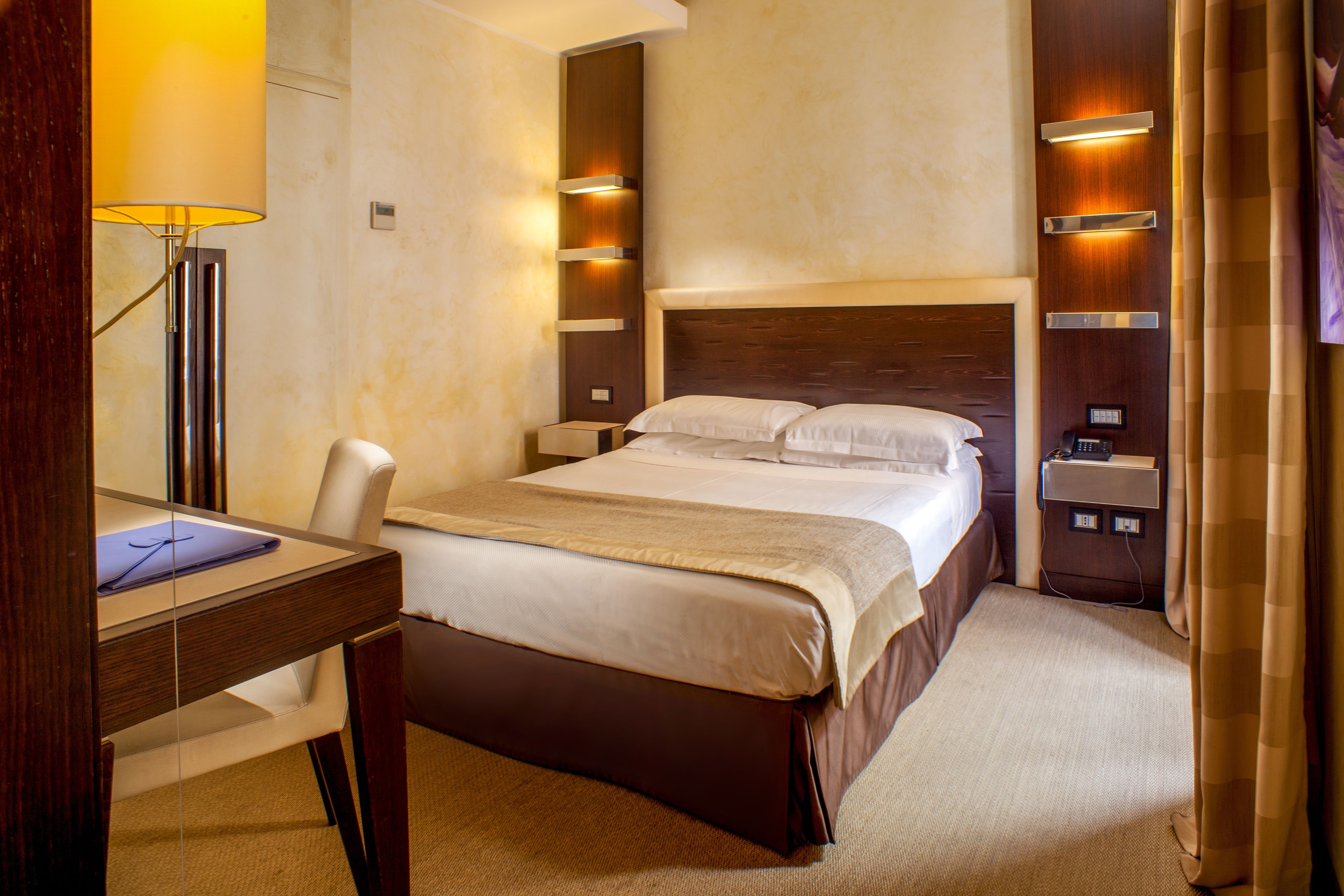 A hotel room with a large bed, bedside tables, a desk with a lamp, and a television.