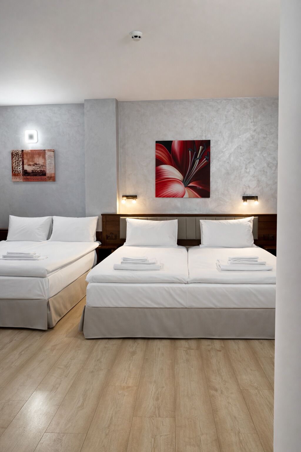 A hotel room with two beds, a light-colored wooden floor, a large floral artwork on the wall, and a ceiling-mounted smoke detector.