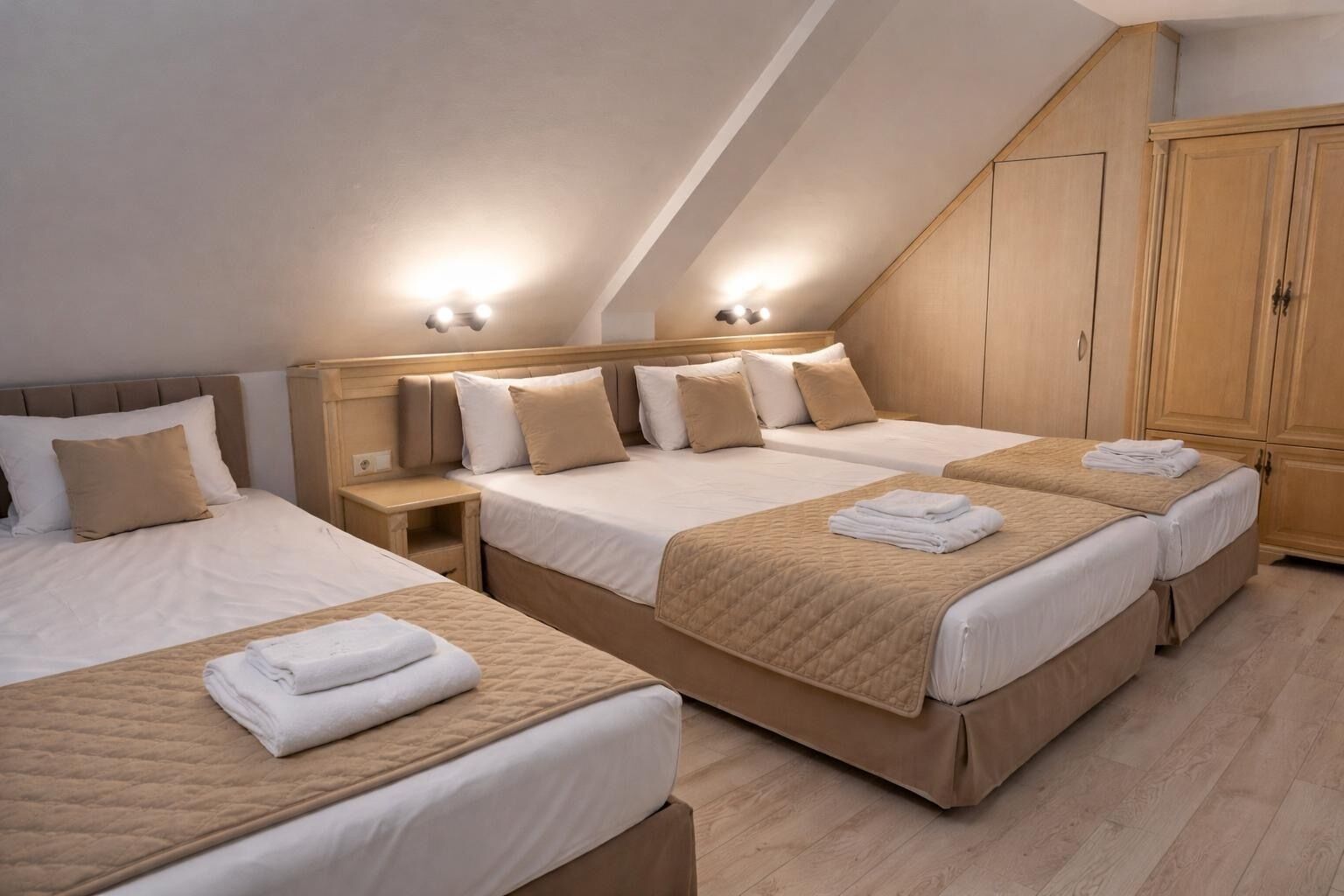 Quarto de hotel com três camas, guarda-roupa de madeira e teto inclinado.