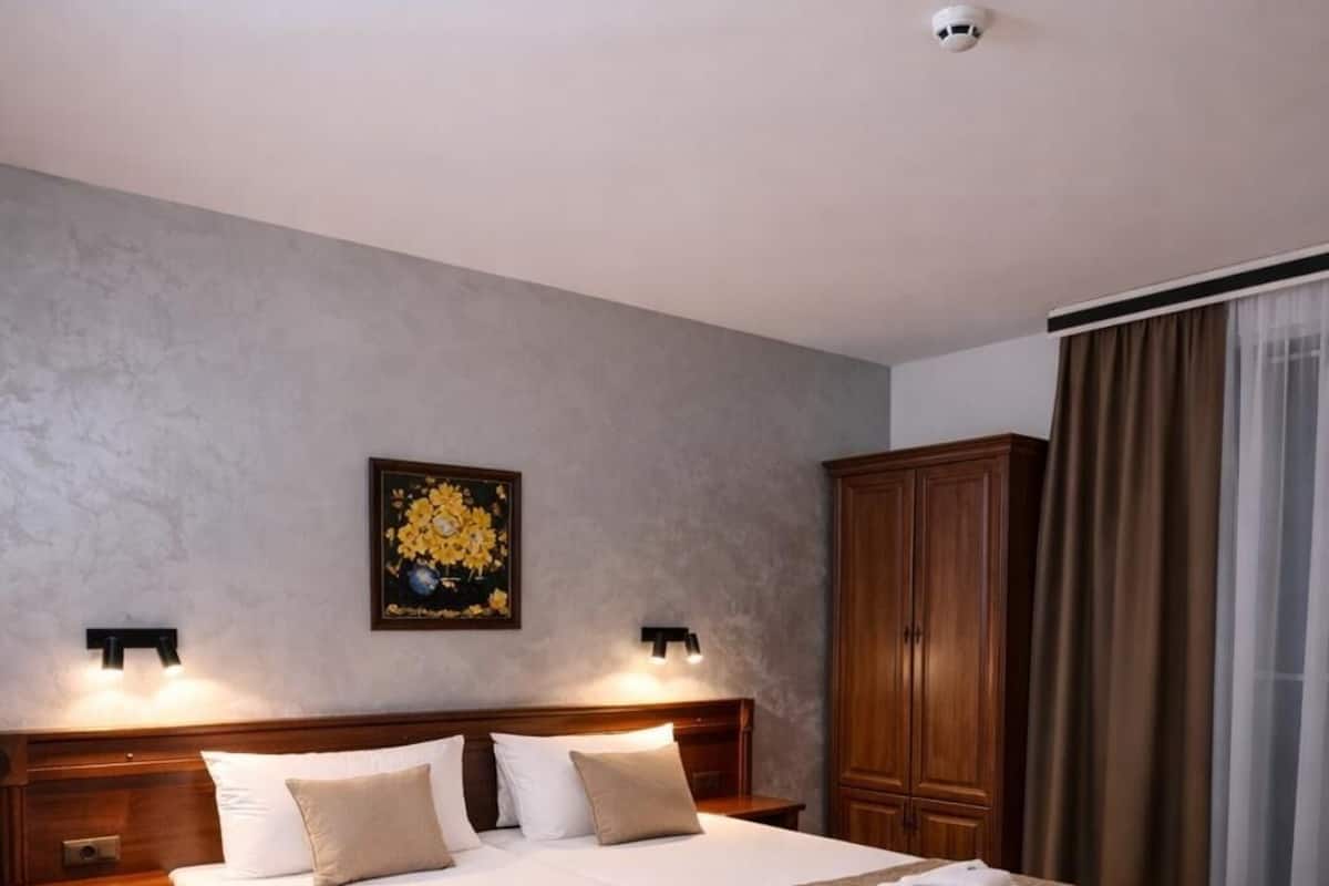 A hotel room with a large bed, wooden headboard, two bedside tables, a nightstand, a wardrobe, and a ceiling-mounted smoke detector.