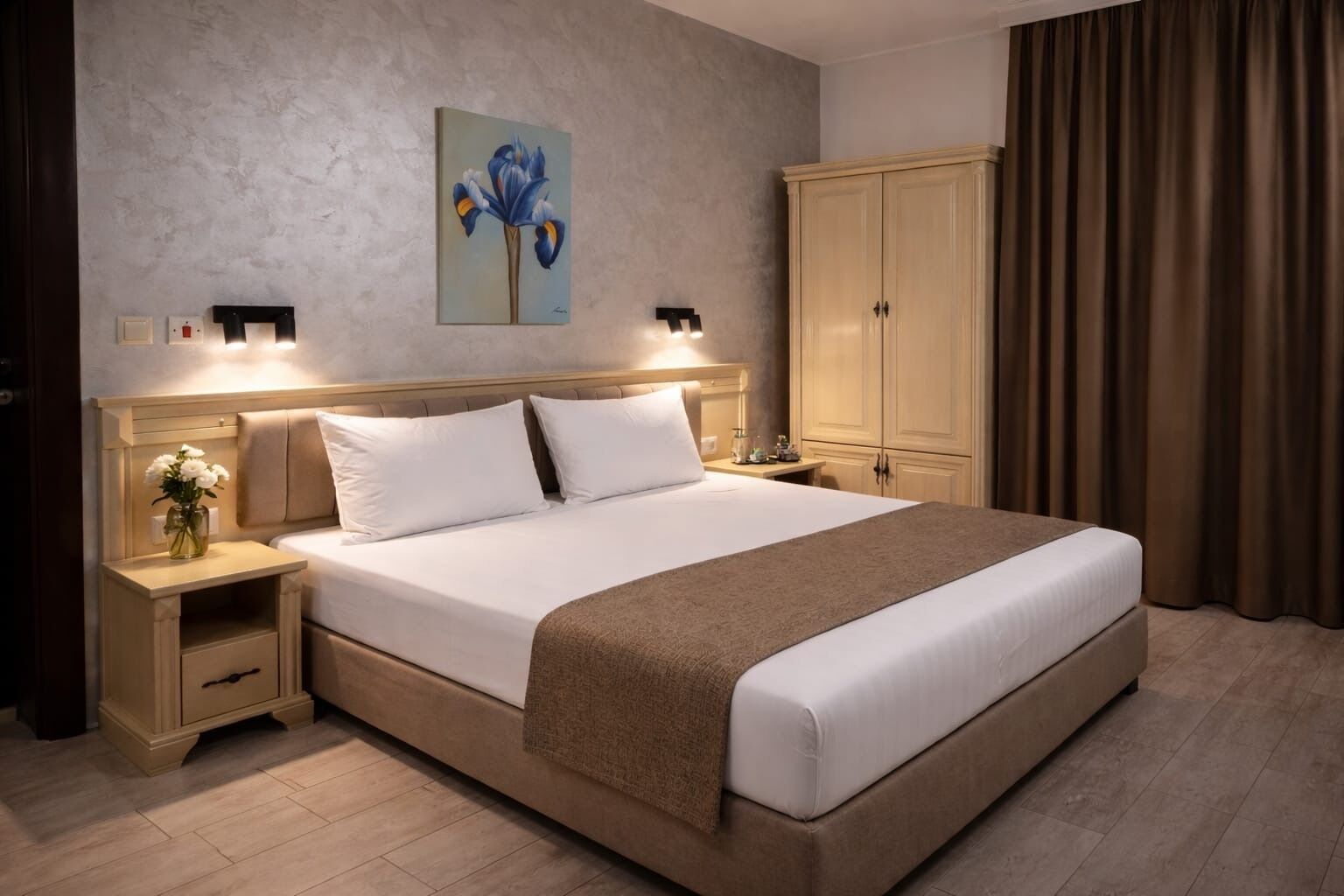 A hotel room with a large bed, bedside tables, a nightstand, a wardrobe, and a painting on the wall.