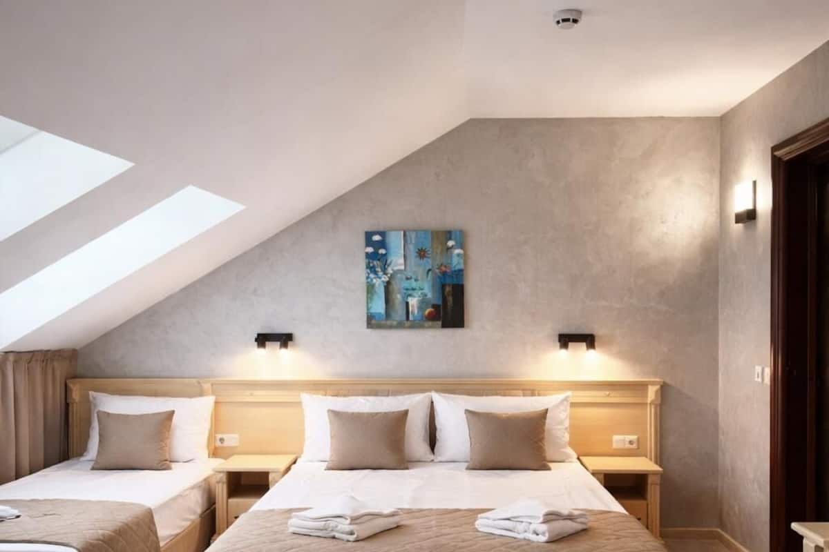A hotel room with a sloped ceiling, a large bed, two smaller beds, a painting on the wall, and a skylight.
