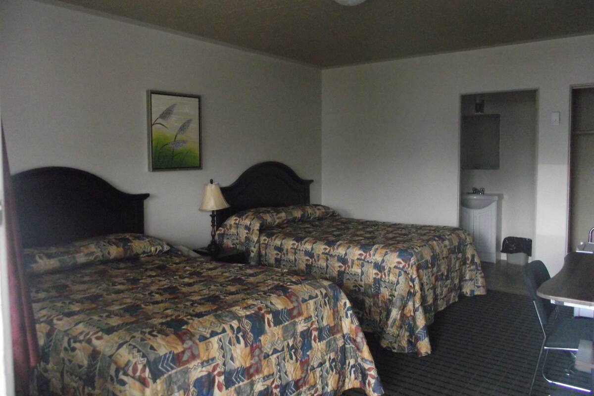 Ett hotellrum med två sängar, ett skrivbord, en stol, en lampa, en tavla och ett badrum.