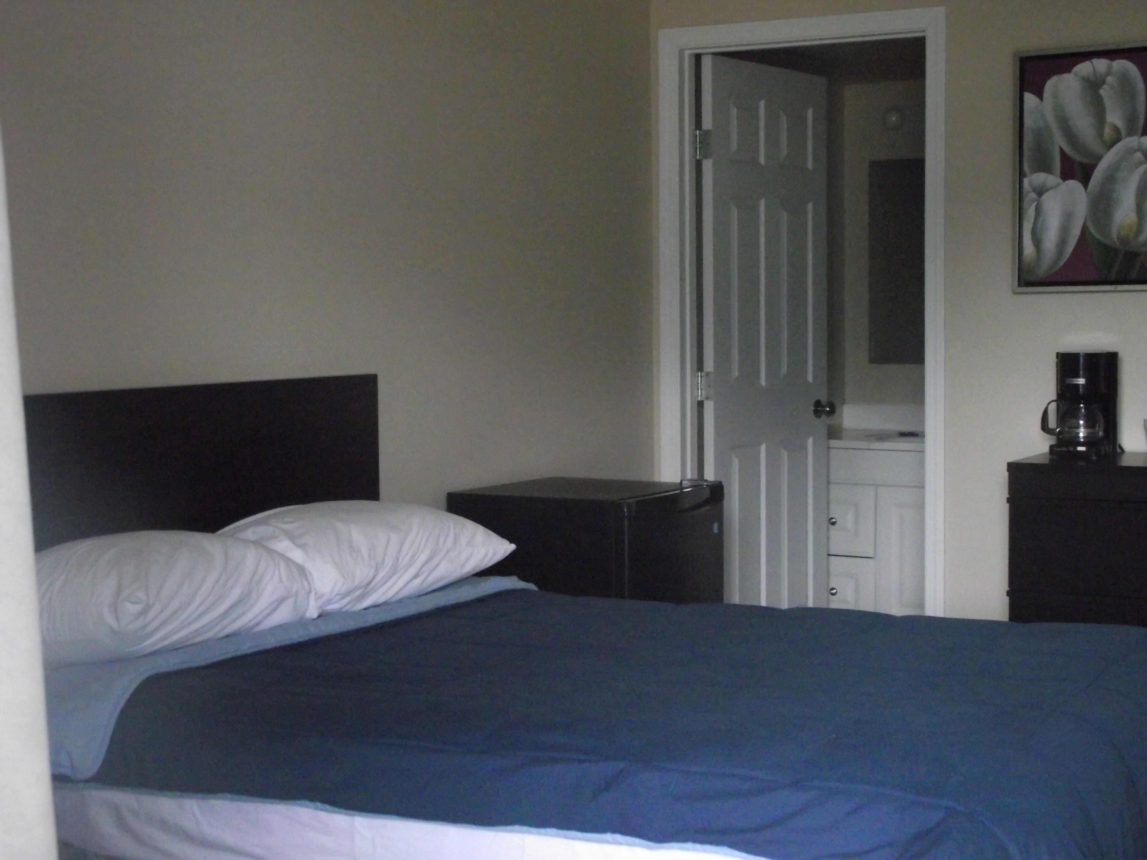 Quarto com cama, mesa de cabeceira, máquina de café e porta para outro cômodo.