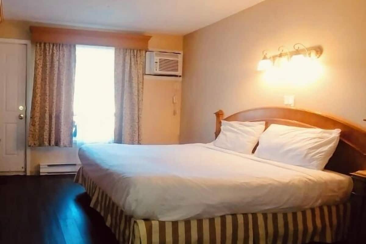 A hotel room with a large bed, a window with curtains, an air conditioning unit, and a wall-mounted light fixture.