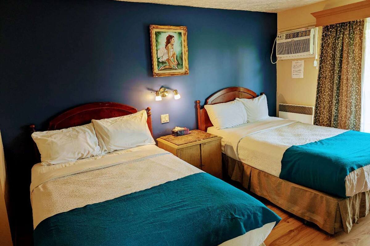 A hotel room with two beds, each with a wooden headboard and white bedding. There is a small bedside table with a lamp, a framed picture on the wall, and an air conditioning unit.