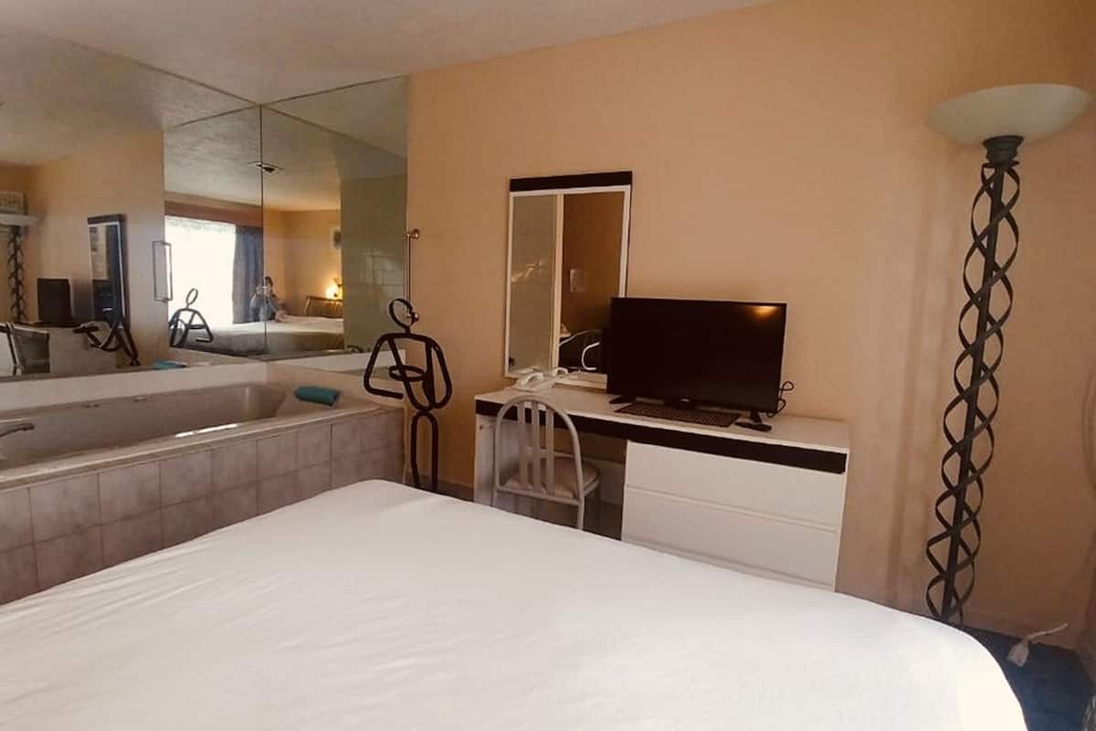 A hotel room with a large bed, a desk with a computer, a TV, a mirror, and a bathroom with a bathtub and sink.