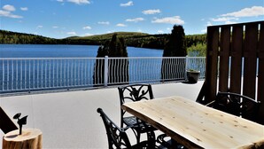 A wooden deck with a table and chairs overlooking a lake, with a railing and trees in the background. - Auberge du Lac Malcom (Sayabec)