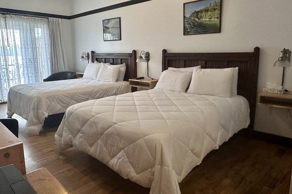 Quarto de hotel com duas camas, cabeceiras de madeira, uma pequena mesa de cabeceira com abajur e uma janela com cortinas.