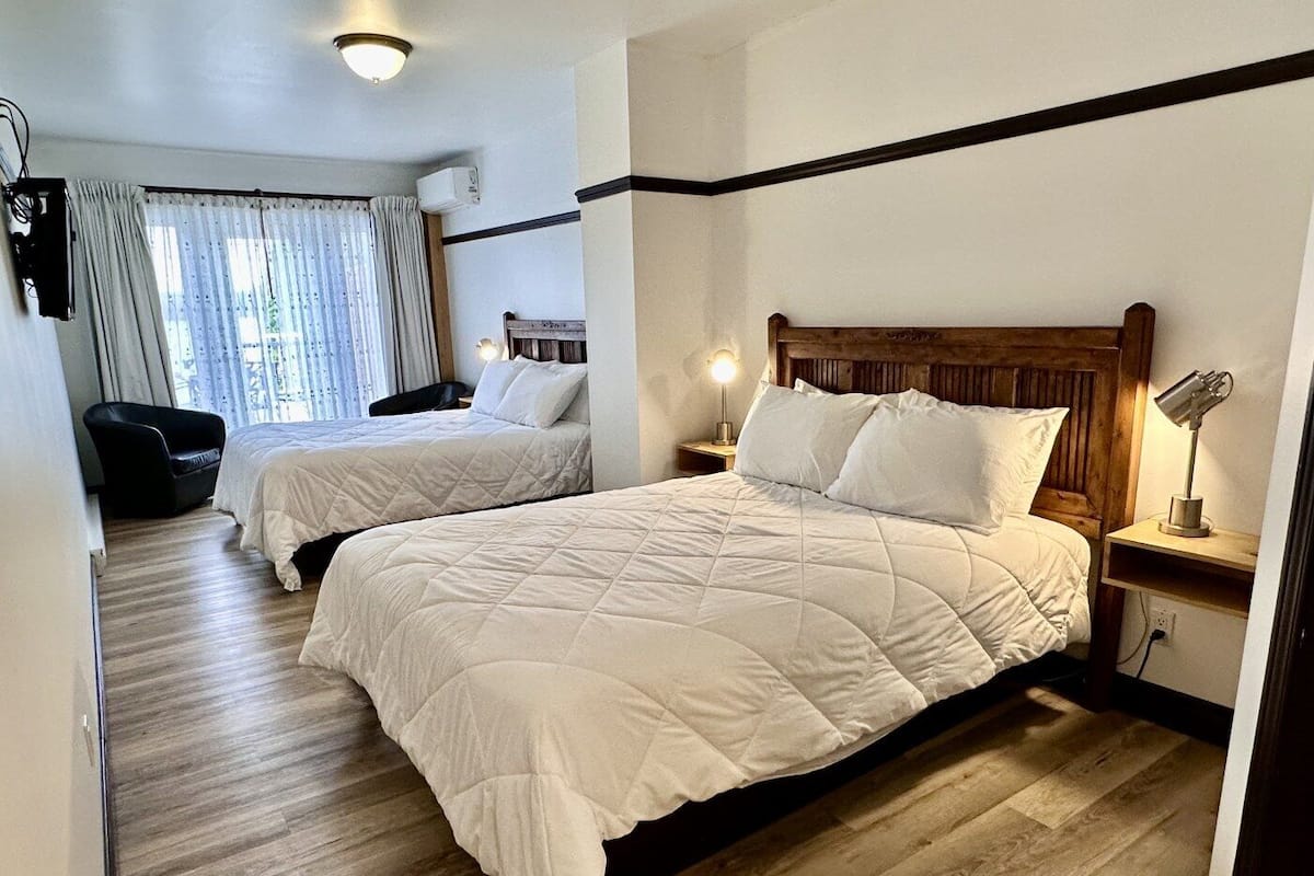 Quarto de hotel com duas camas, uma cabeceira de madeira, uma mesa de cabeceira com abajur, uma cadeira e uma janela com cortinas.
