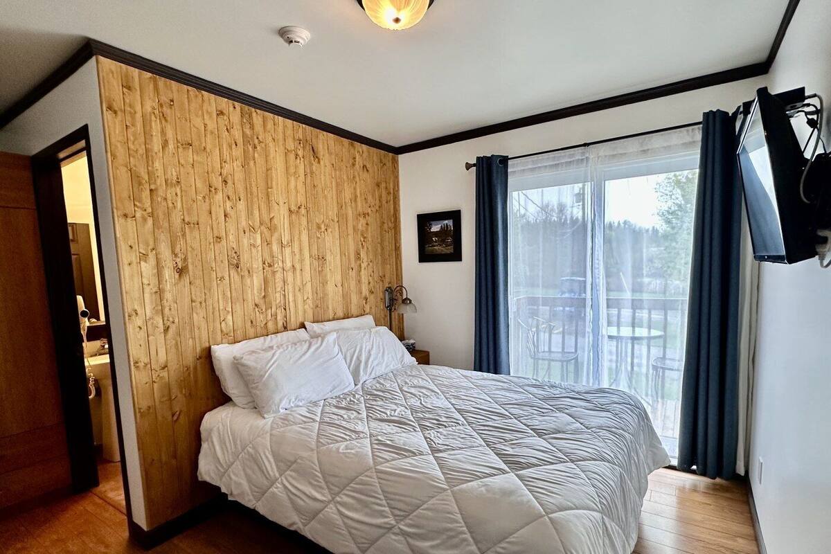Quarto com cabeceira de madeira, cama com edredom branco acolchoado, mesa de cabeceira pequena, luminária fixada na parede e janela com cortinas.
