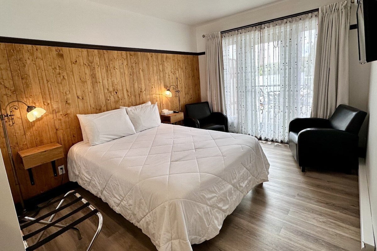 Quarto de hotel com cabeceira de madeira, cama grande com roupa de cama branca, duas cadeiras pretas, mesa com luminária e janela com cortinas.