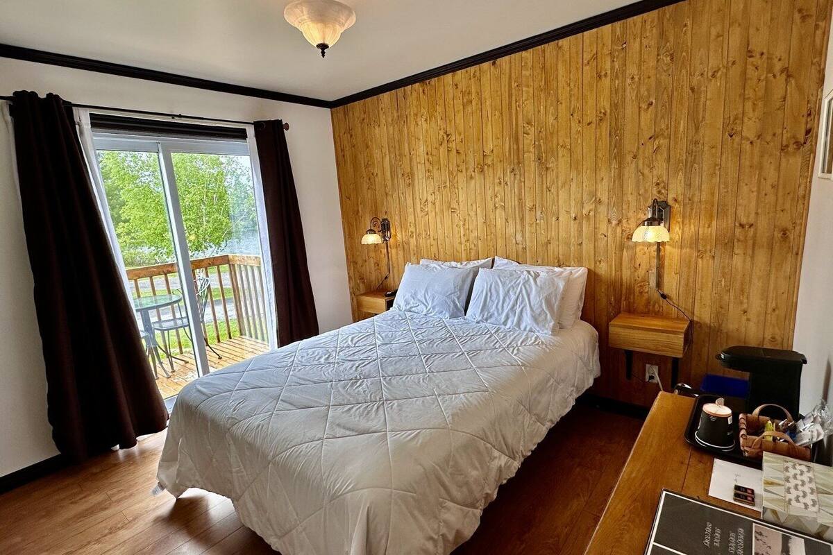 Quarto com cama, paredes de painel de madeira, janela com cortinas, deck de madeira na parte externa e duas luminárias de cabeceira.