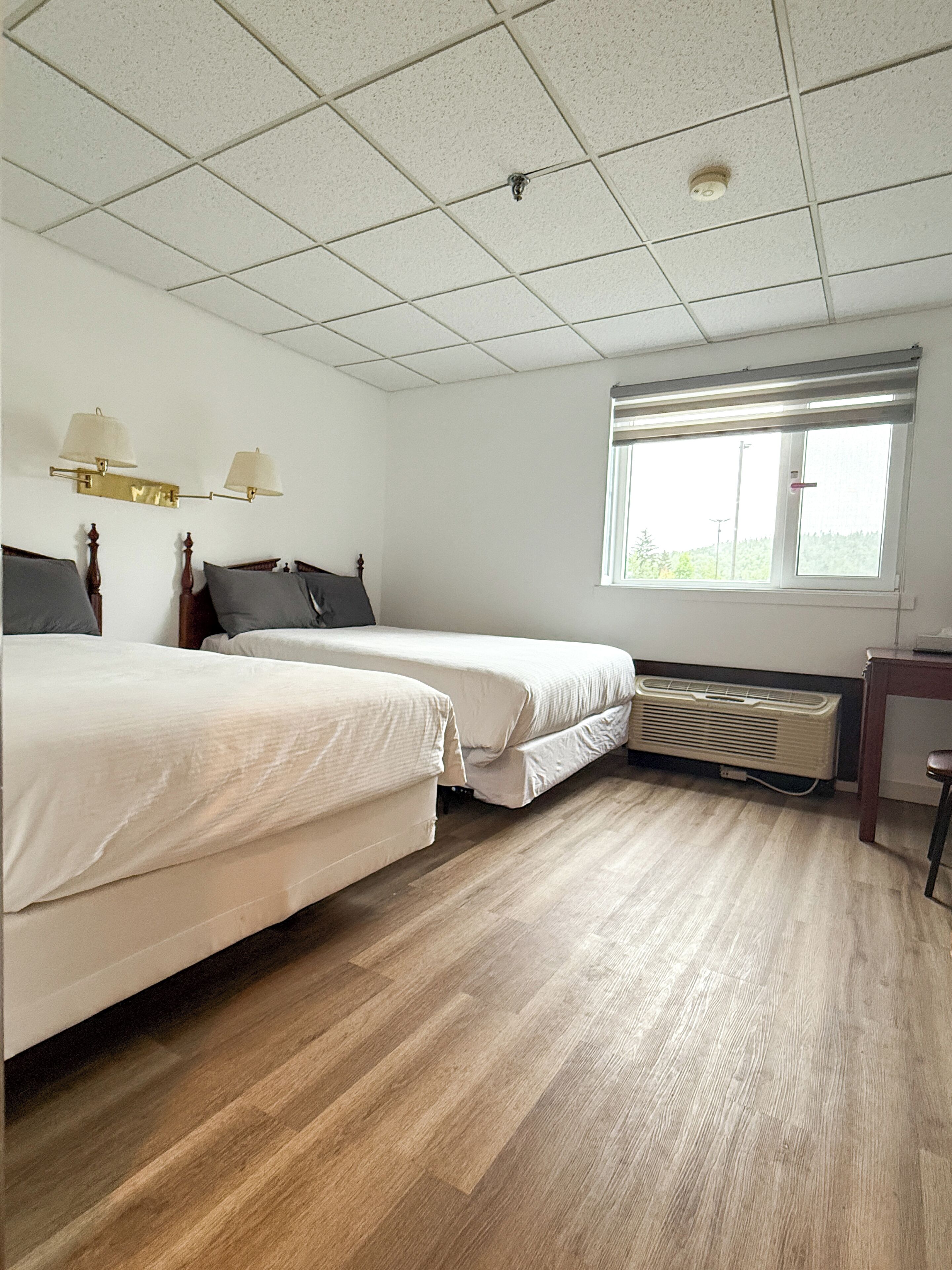 A hotel room with two beds, a window, a heater, and a small table.
