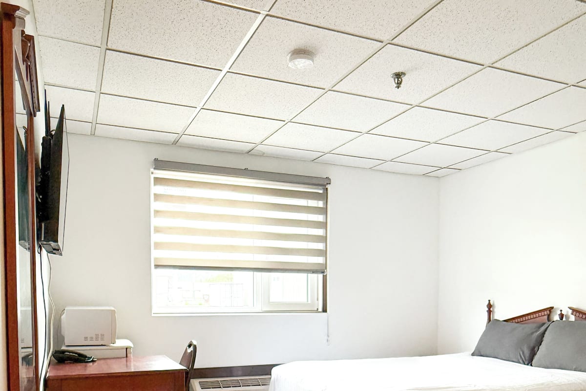 A bedroom with a bed, a desk, a chair, a radiator, and a window with blinds.