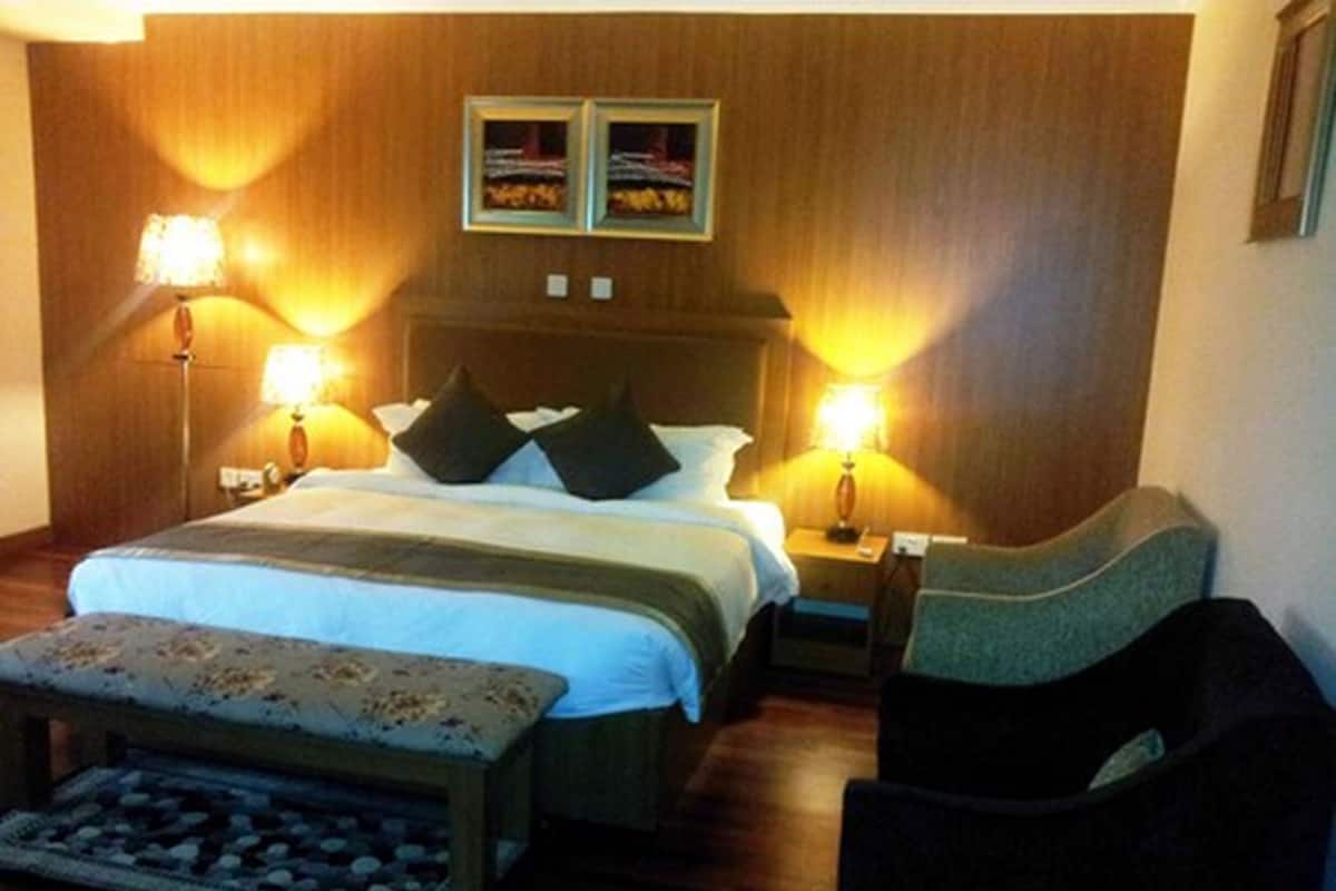 Una habitación de hotel con una cama grande, dos lámparas de noche, una mesita pequeña, una silla y dos cuadros en la pared.