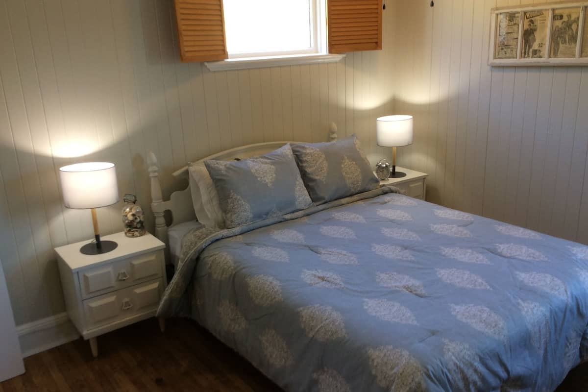 Un lit bien fait, recouvert d’une couvre-lit bleue à motifs et agrémenté d’oreillers, avec de chaque côté des tables de chevet sur lesquelles sont posées des lampes et un petit objet décoratif.
