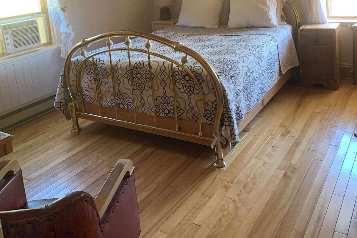 Une chambre à coucher avec un lit en bois, une table de chevet avec une lampe, une fenêtre avec des rideaux et un tableau encadré au mur.