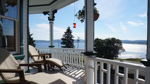 A wooden deck with white railings and beige cushioned chairs, overlooking a body of water and distant mountains. - B&B Le Manoir du vieux Presbytère (Port-Daniel-Gascons)