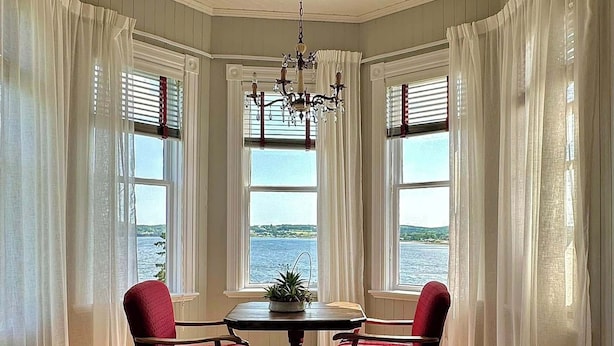 A room with a bay window overlooking a body of water, a chandelier, and two red chairs around a table. - B&B Le Manoir du vieux Presbytère (Port-Daniel-Gascons)