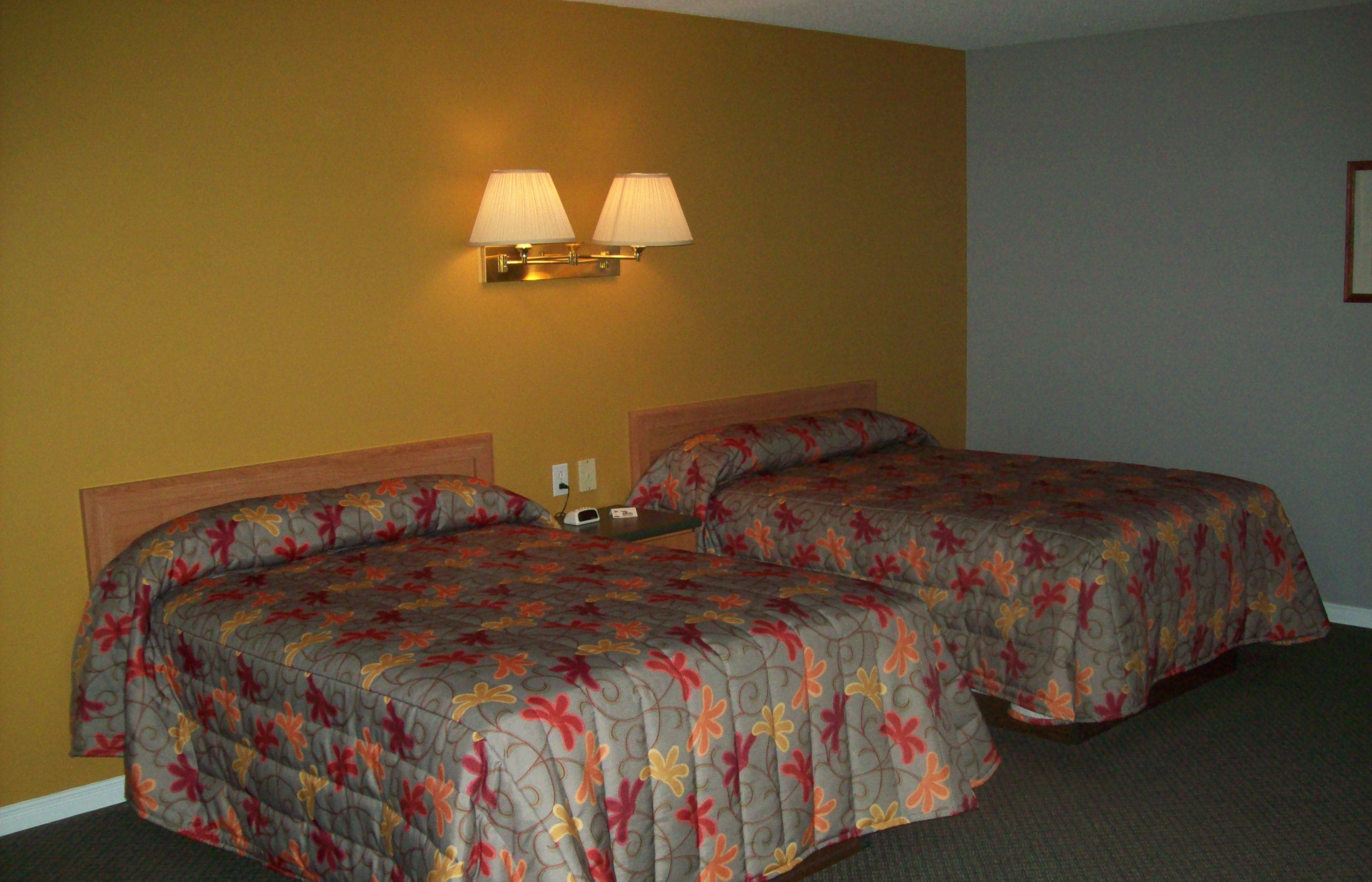 A hotel room with two beds, each with a floral-patterned bedspread, a wooden headboard, and a wall-mounted lamp.