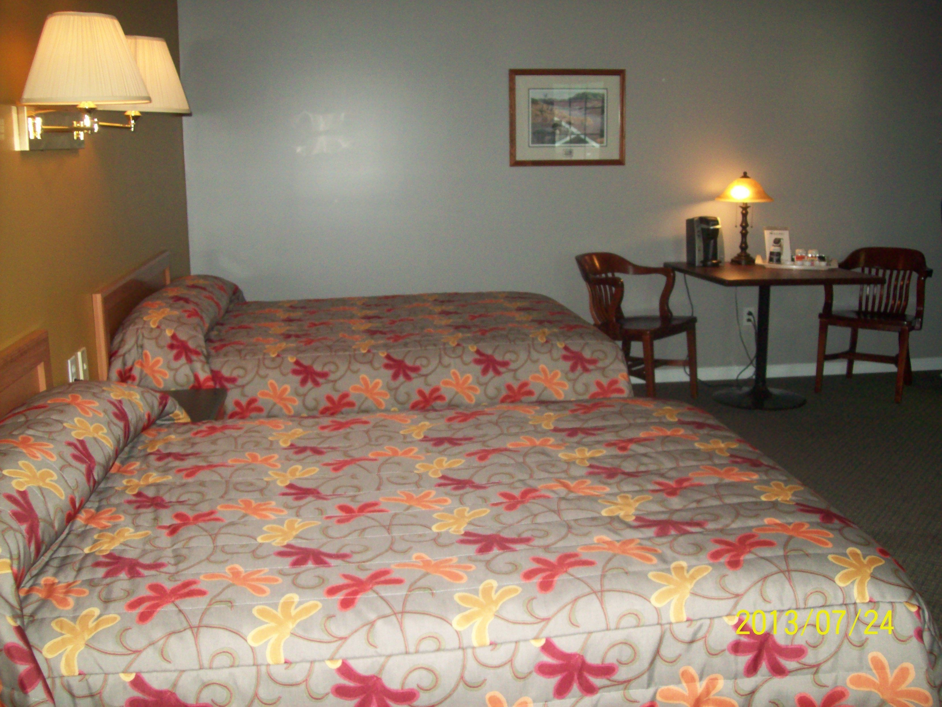 A hotel room with two beds, a desk, two chairs, a lamp, and a framed picture on the wall.
