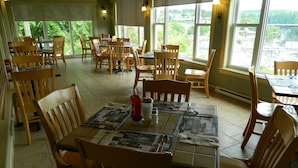 A restaurant with multiple wooden tables and chairs, large windows with a view of greenery and buildings, and a tiled floor. - Hill Top Motel & Restaurant (Grand Falls)