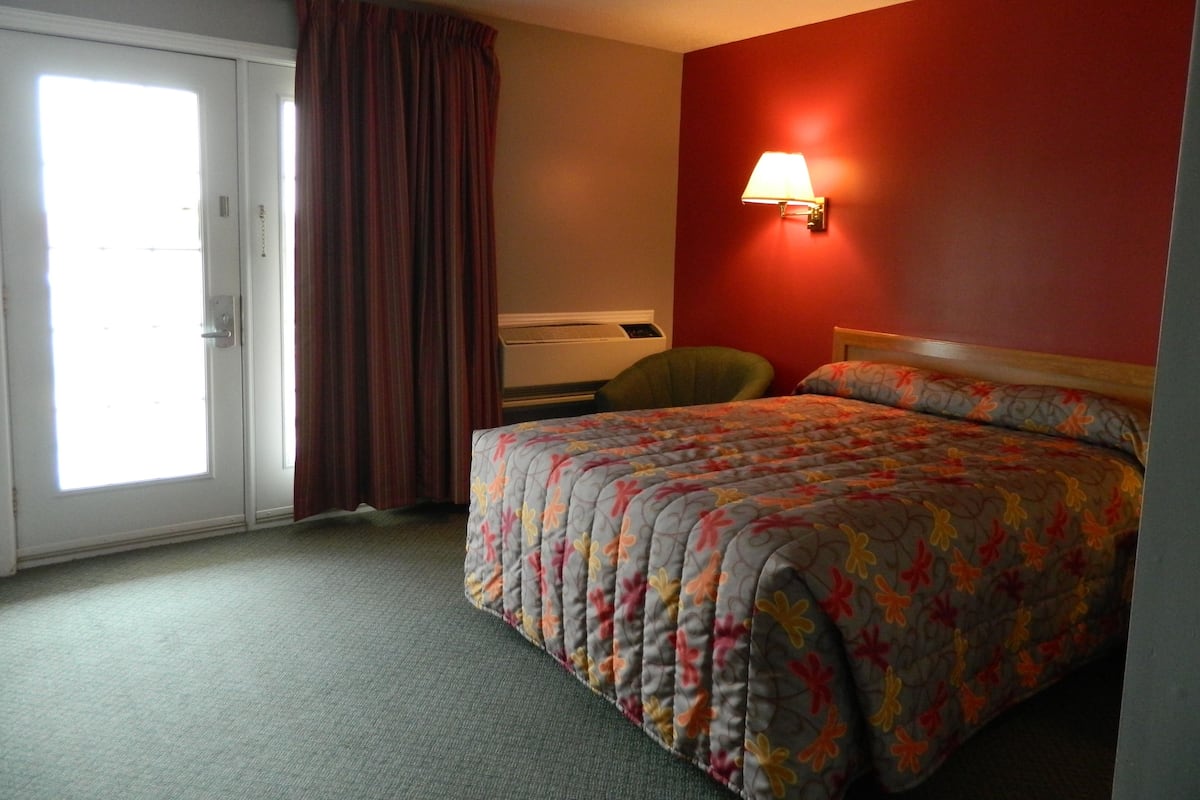 Una habitación de hotel con cama, escritorio, silla, ventana con cortinas y una lámpara fijada a la pared.