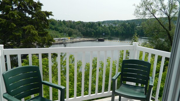 A balcony with two green chairs, a white railing, and a view of a river and forested area. - Hill Top Motel & Restaurant (Grand Falls)