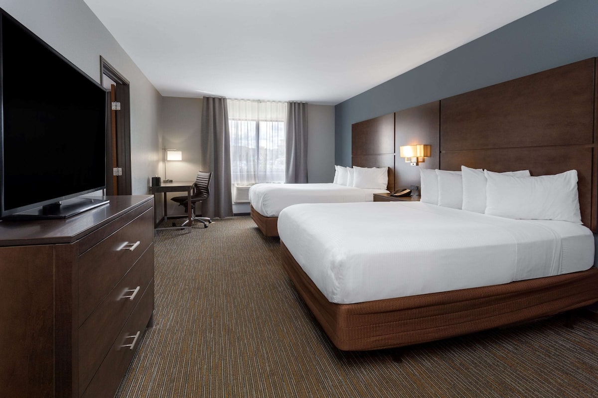 A hotel room with two beds, a desk, a TV, and a large window with curtains.
