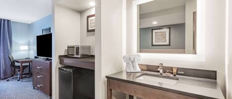 A hotel room with a desk, chair, minibar, and a large mirror above the sink.