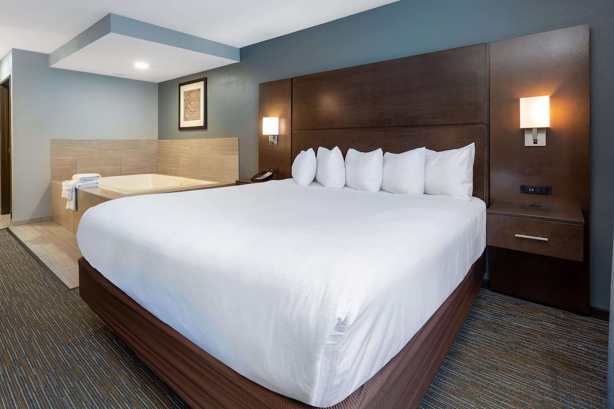 A large bed with a wooden headboard, a bedside table with a lamp, and a small bathroom with a bathtub and towels.