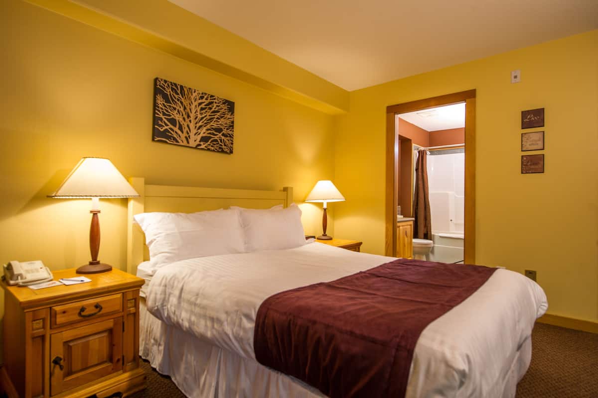 A hotel room with a bed, two bedside lamps, a nightstand with a phone, a painting on the wall, and a bathroom door.