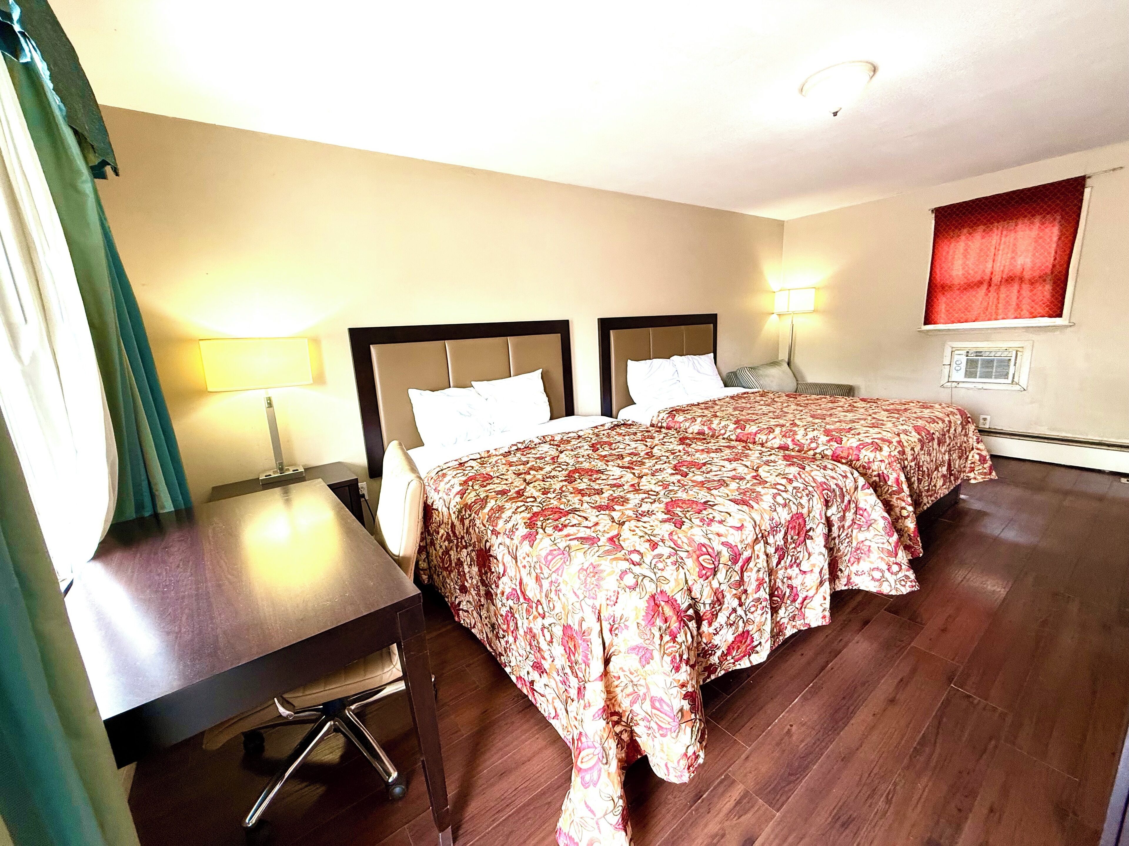 A hotel room with two beds, a desk, a lamp, and a window with curtains.