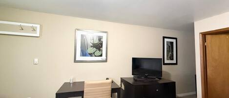 Une petite pièce bien éclairée, avec un bureau, une chaise et un téléviseur posé sur un meuble.