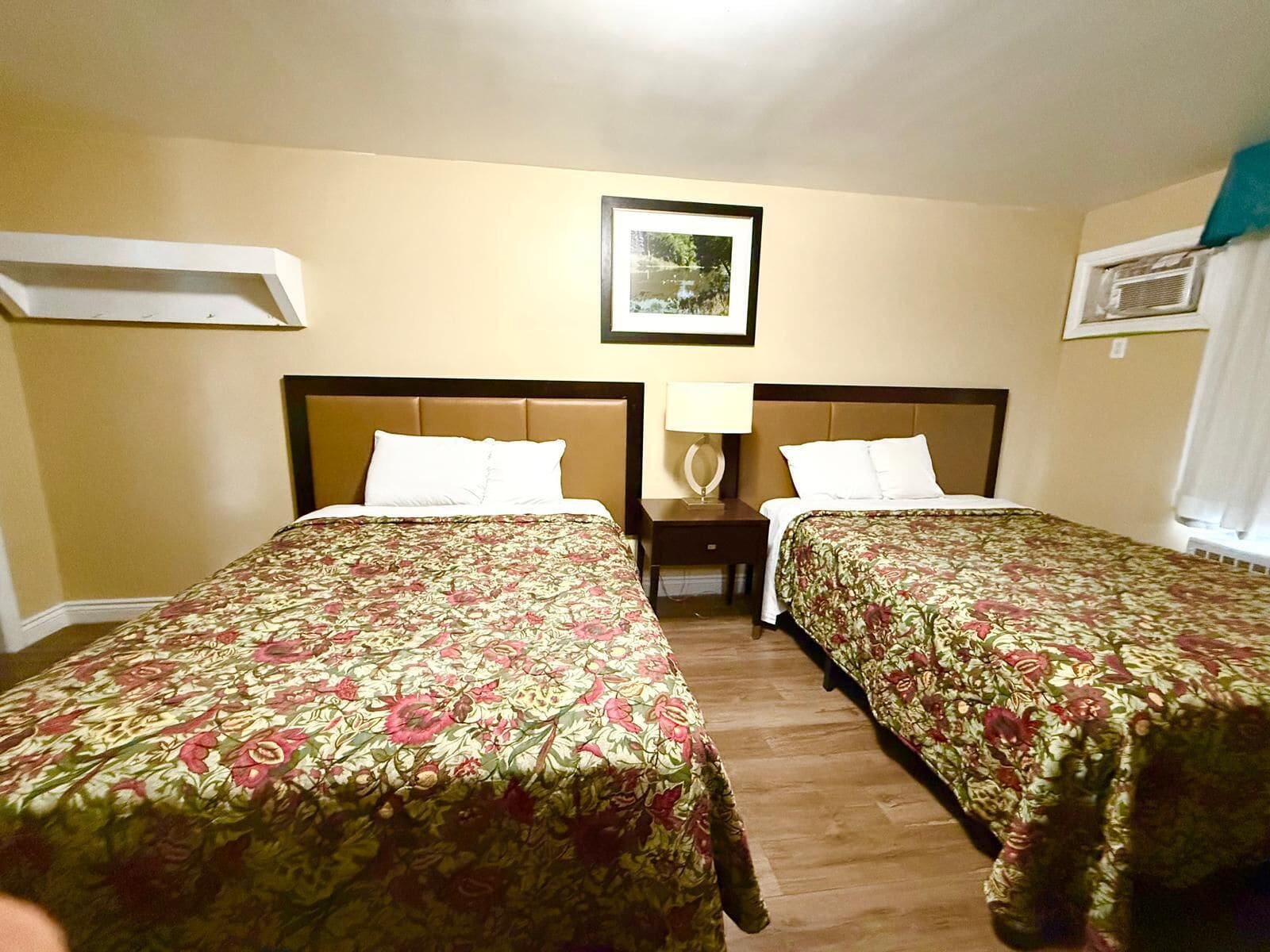 Habitación de hotel con dos camas, cada una con cubrecamas florales, una mesita de noche con lámpara y un cuadro en la pared.