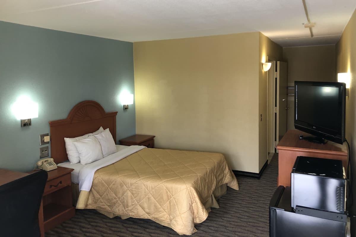 A hotel room with a bed, bedside table, lamp, TV, and a desk with a phone.