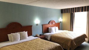 Quarto de hotel com duas camas, cada uma com cabeceira de madeira, roupa de cama branca e edredom acolchoado bege. Há uma mesa de cabeceira com telefone e luminária fixa na parede.