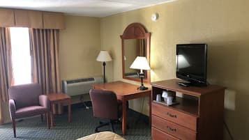 Una habitación de hotel con cama, escritorio y silla, televisor, lámpara, espejo y calefactor.