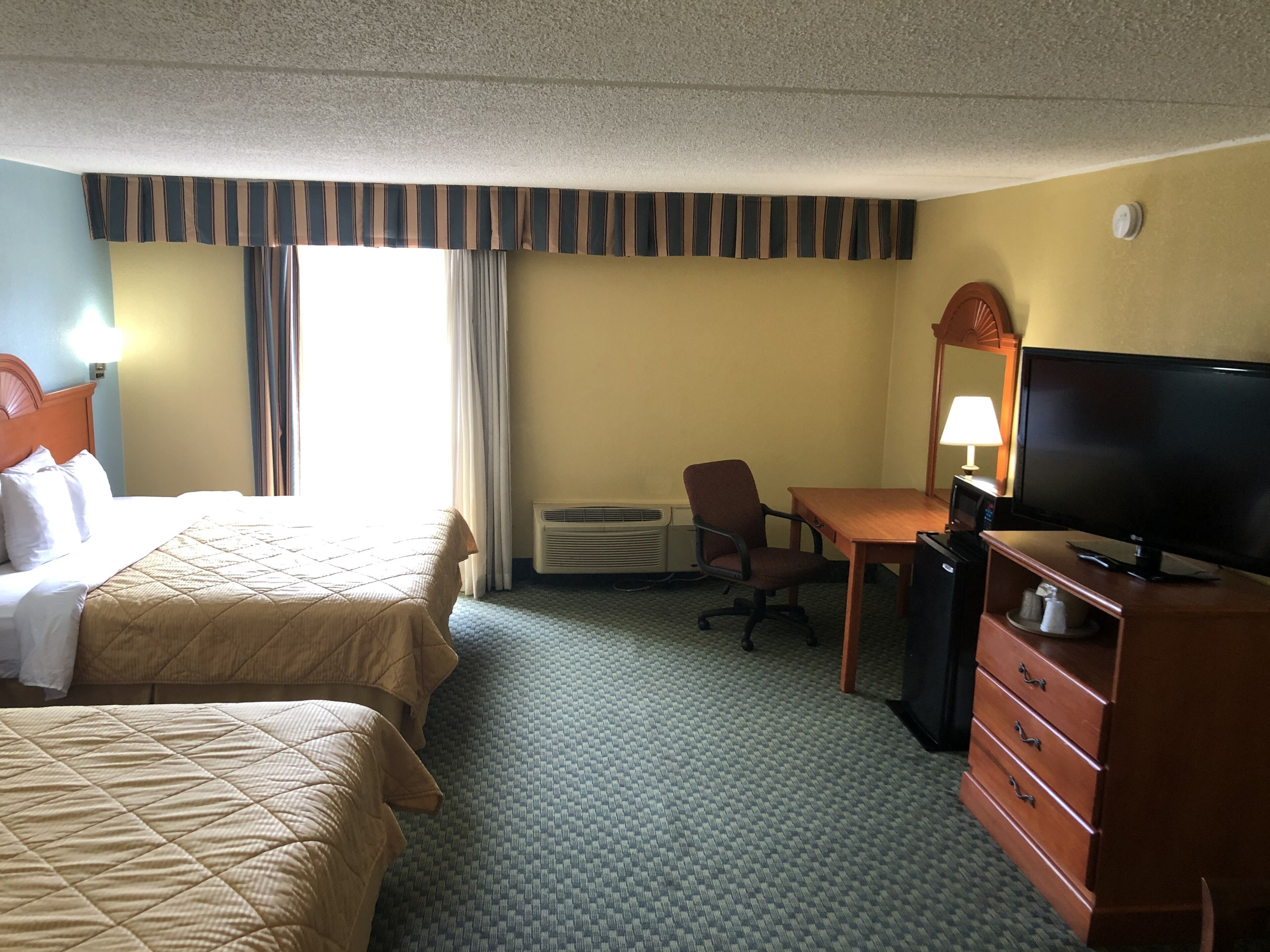 A hotel room with two beds, a desk, a television, a dresser, and an air conditioning unit.
