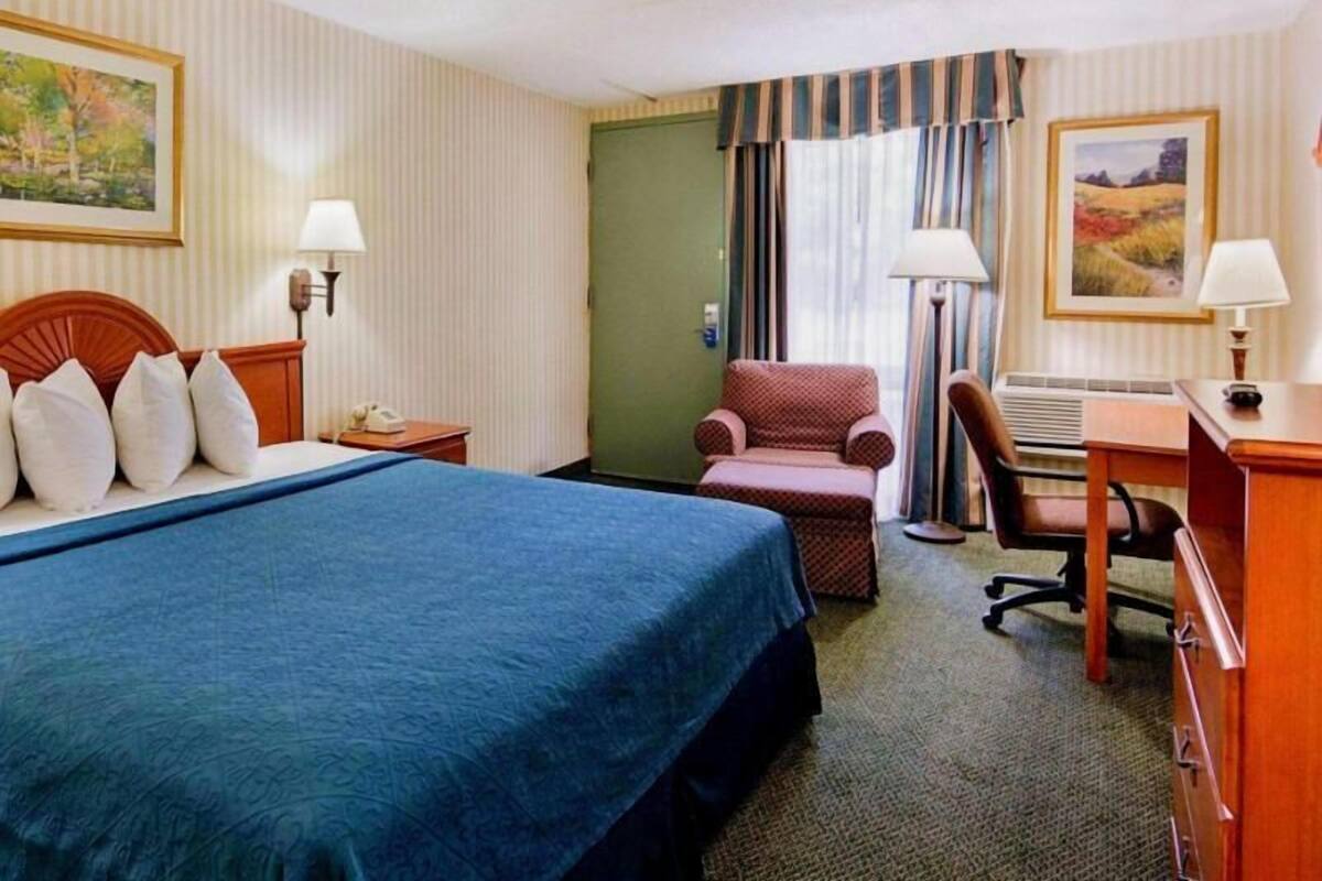 A hotel room with a bed, a desk, a chair, a lamp, and a painting on the wall.