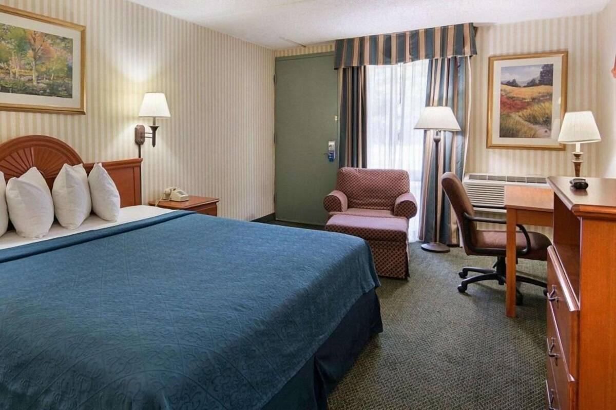 A hotel room with a bed, a desk, a chair, a lamp, a telephone, and a door.