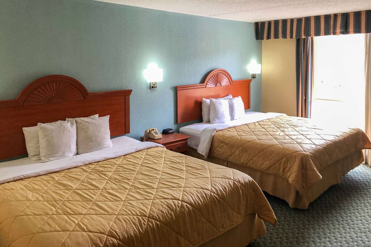 A hotel room with two beds, each with a wooden headboard and a quilted bedspread. There is a nightstand with a telephone and a lamp between the beds.