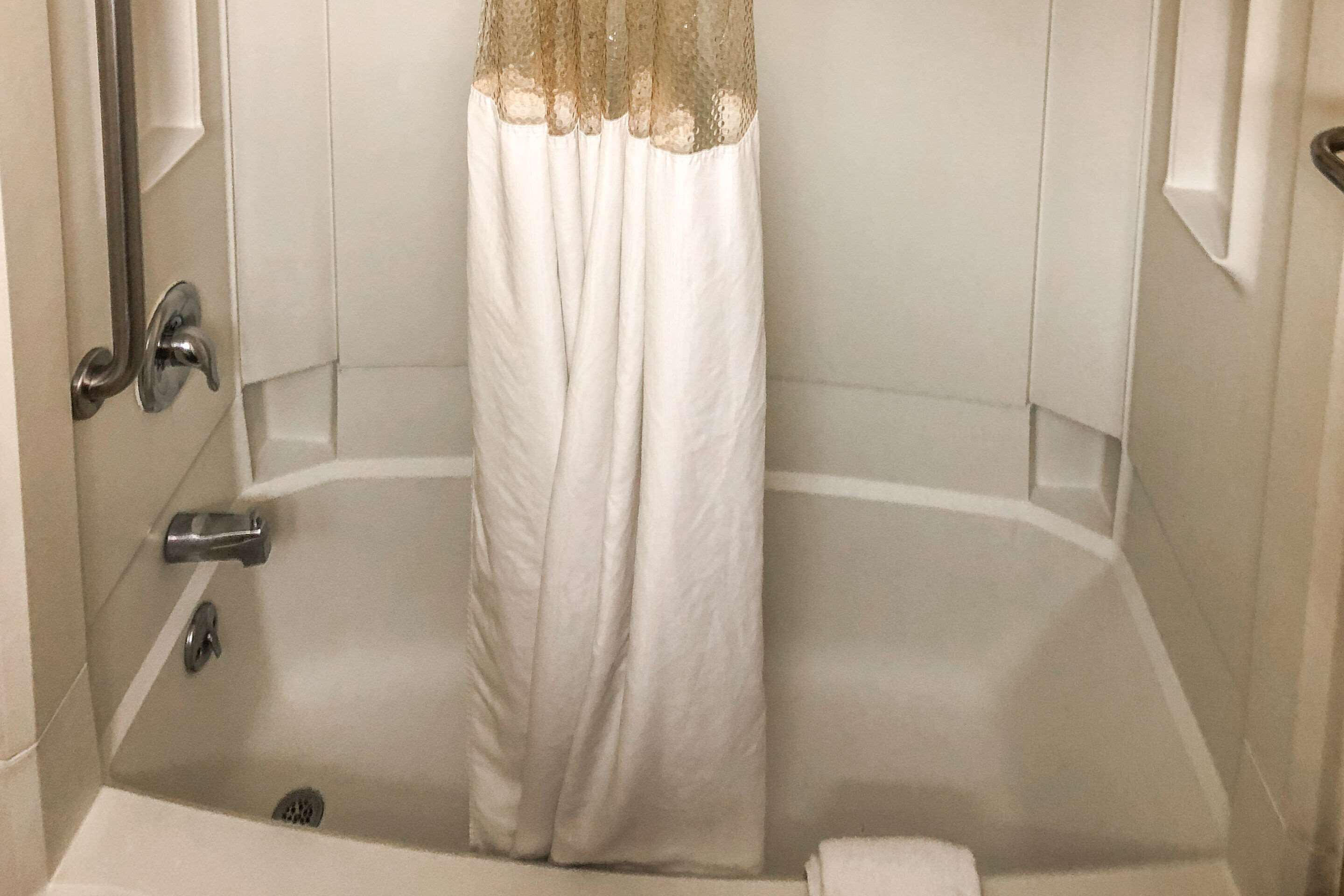 Una bañera con una cortina blanca, un cabezal de ducha manual y una toalla en el suelo.