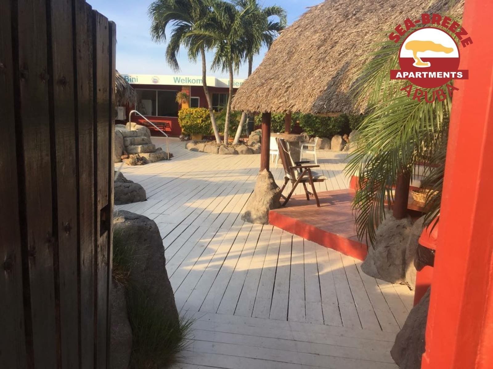 A wooden deck with a thatched-roof structure, palm trees, and a sign for 'Sea Breeze Apartments' in the background.