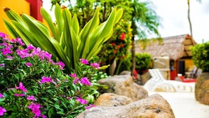 A garden with green foliage, pink flowers, and a thatched-roof structure. - Sea Breeze Apartments (Savaneta)