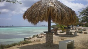 A beach with a thatched umbrella, a stone pillar, and a bench. - Sea Breeze Apartments (Savaneta)