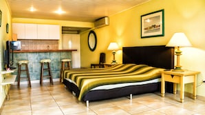A compact hotel room with a bed, two bedside lamps, a small table, a chair, a stool, a TV, and a kitchen area with a counter and stools. - Sea Breeze Apartments (Savaneta)